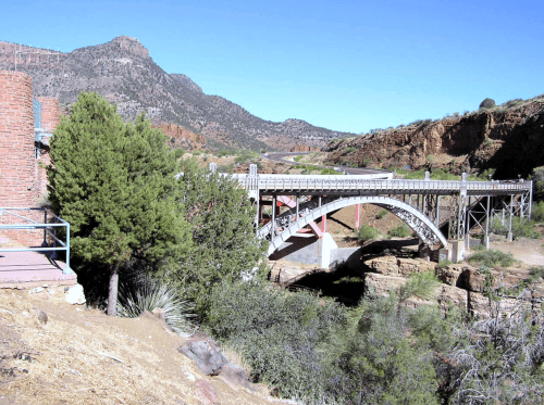 A scenic view of a bridge spanning a canyon, surrounded by mountains and greenery under a clear blue sky.